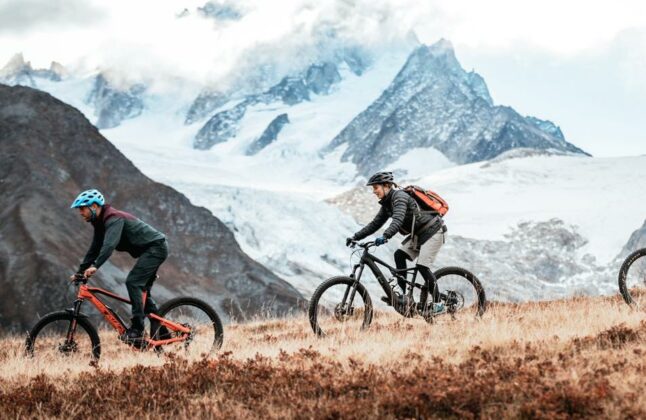 Two mountain bikers riding a trail with snow-capped Alpine peaks in the distance, during their Haute Route traverse.