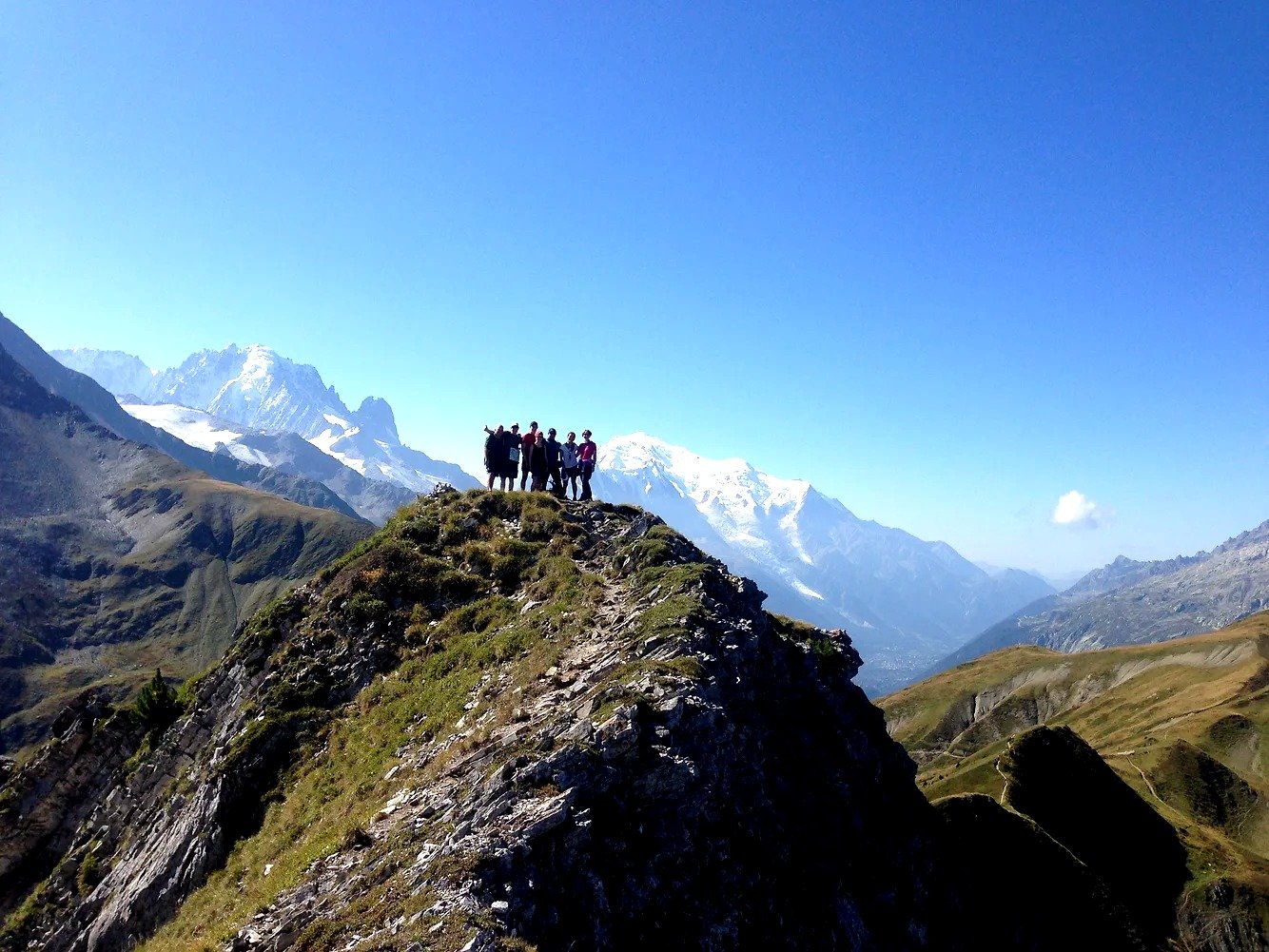A group of TMB hikers posing for a photo standing on a ridge surrounded by snow-capped mountains in the distance.