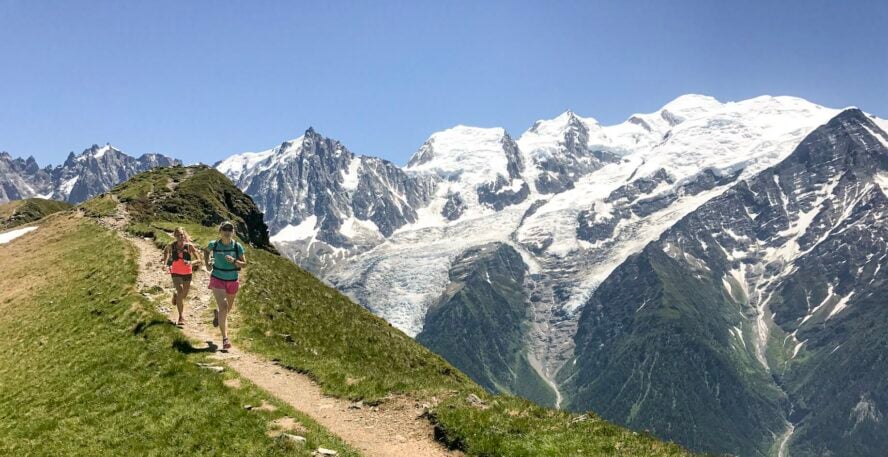 Two women running on a dirt ridgeline trail in the Alps with large snow-capped mountains in the background.