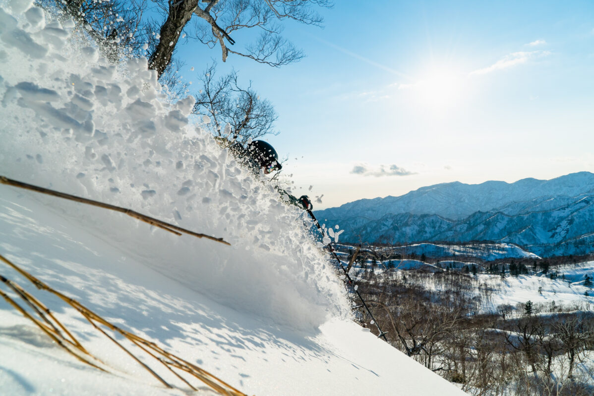 A skier in a cloud of snow with mountains and the sun in the background.
