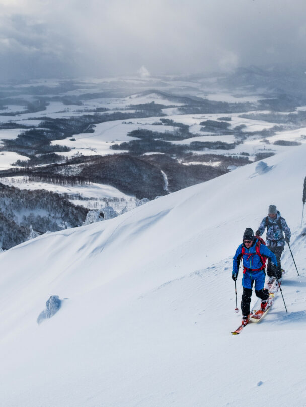 Three people going uphill on a ski tour with fog and snow in the background.