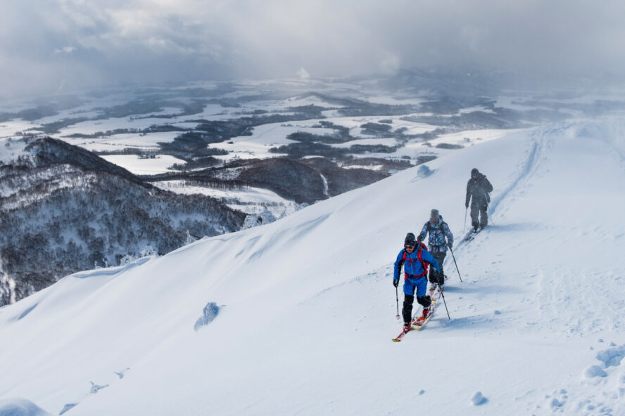 Three people going uphill on a ski tour with fog and snow in the background.