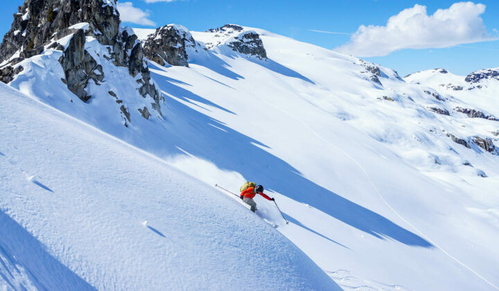 A person in a red jacket and yellow backpack skiing down a mountain face with no other tracks, rocky peaks in the background, and clear skies.