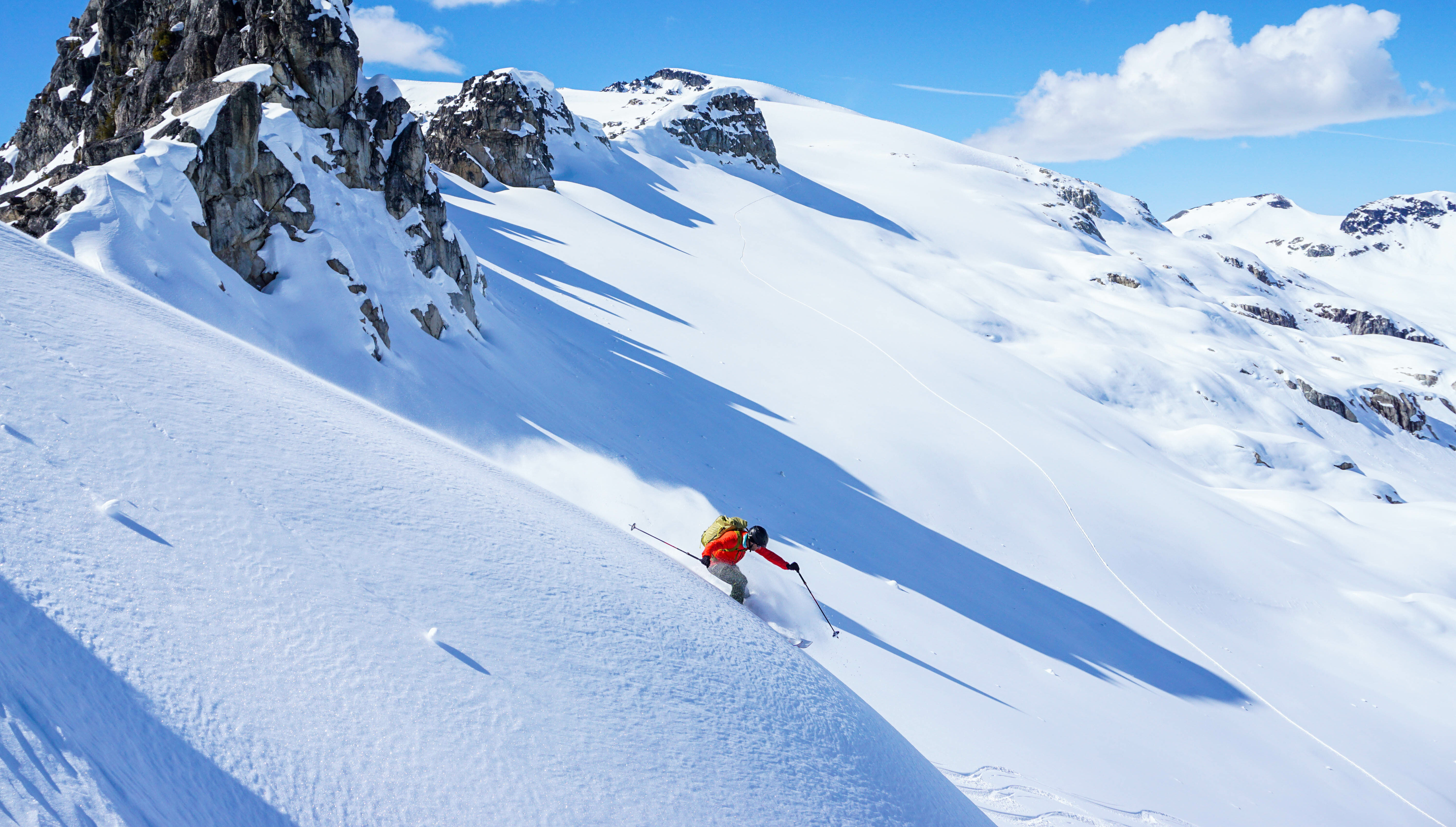 A person in a red jacket and yellow backpack skiing down a mountain face with no other tracks, rocky peaks in the background, and clear skies.