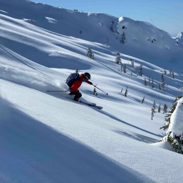 Person with a red jacket skiing down the face of a mountain on untouched snow.