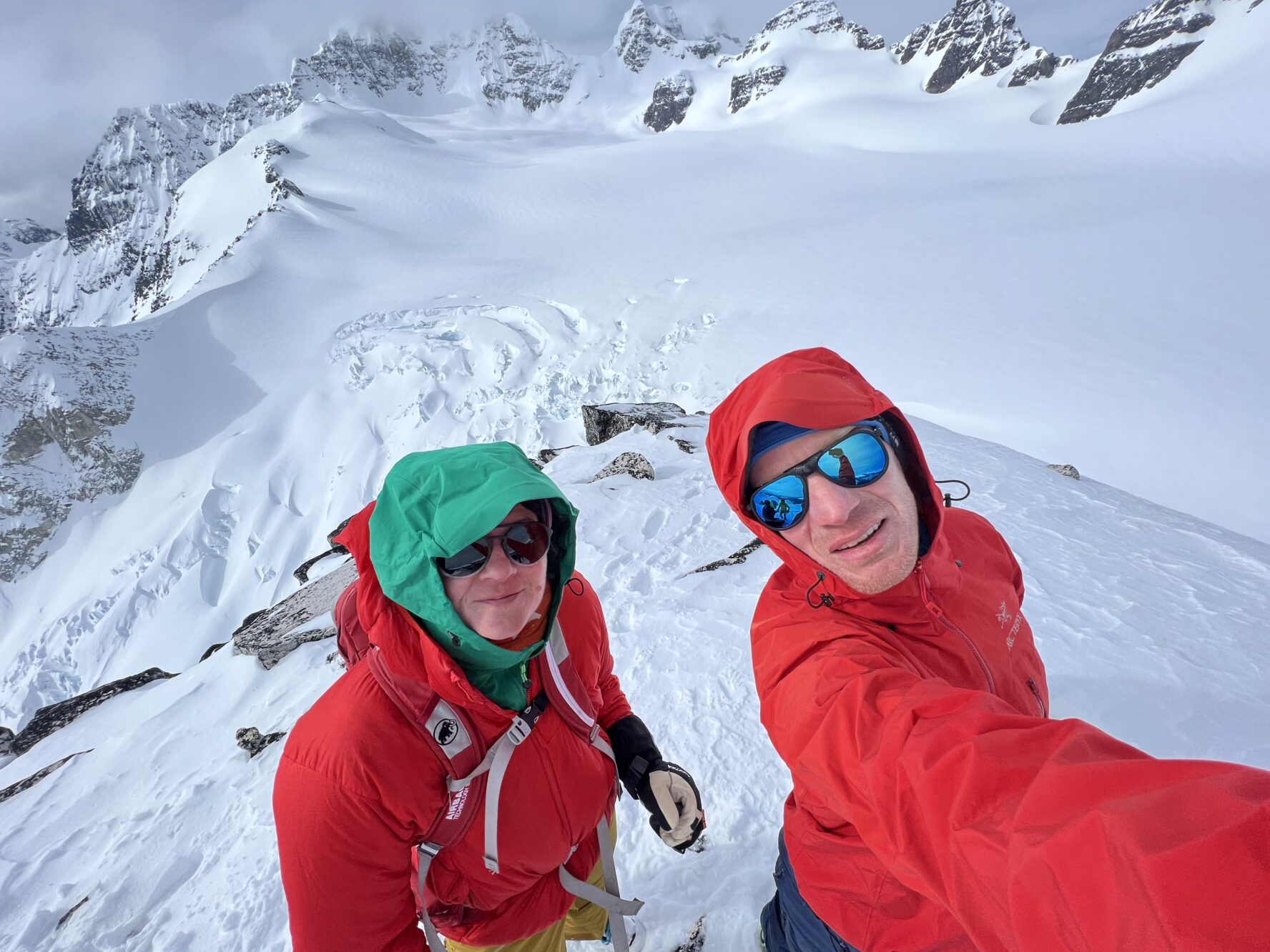 A couple in red jackets taking a selfie on top of a mountain after a backcountry ski tour.