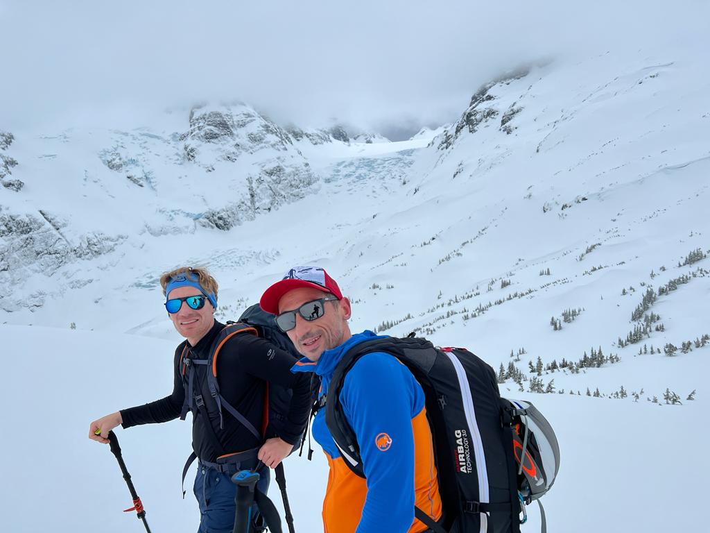 Two people posing for a photo on a ski tour in British Columbia with the peaks behind them covered in snow.