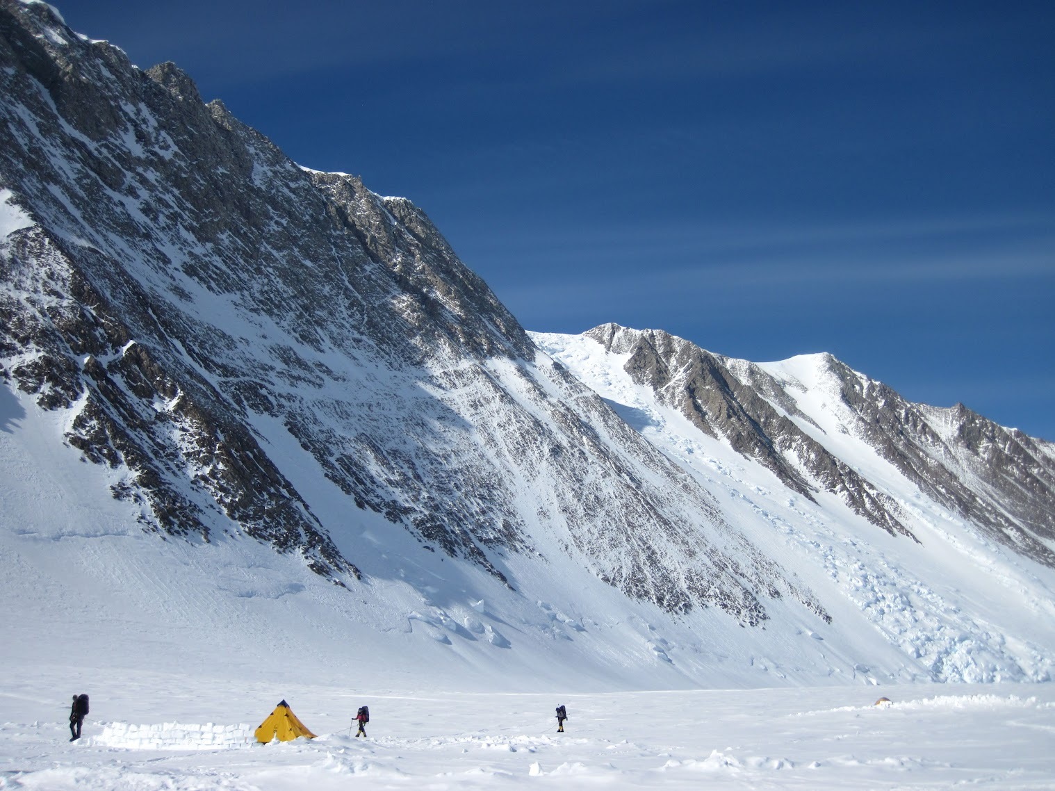 A valley in Antarctica, surrounded by rugged mountains, with two tents and mountaineers seen from a distance.