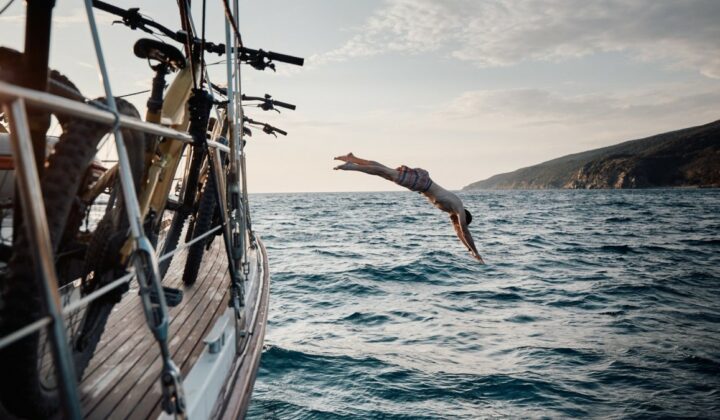 A mountain biker jumping into the sea from a sailboat with e-mountain bikes heading to Elba.