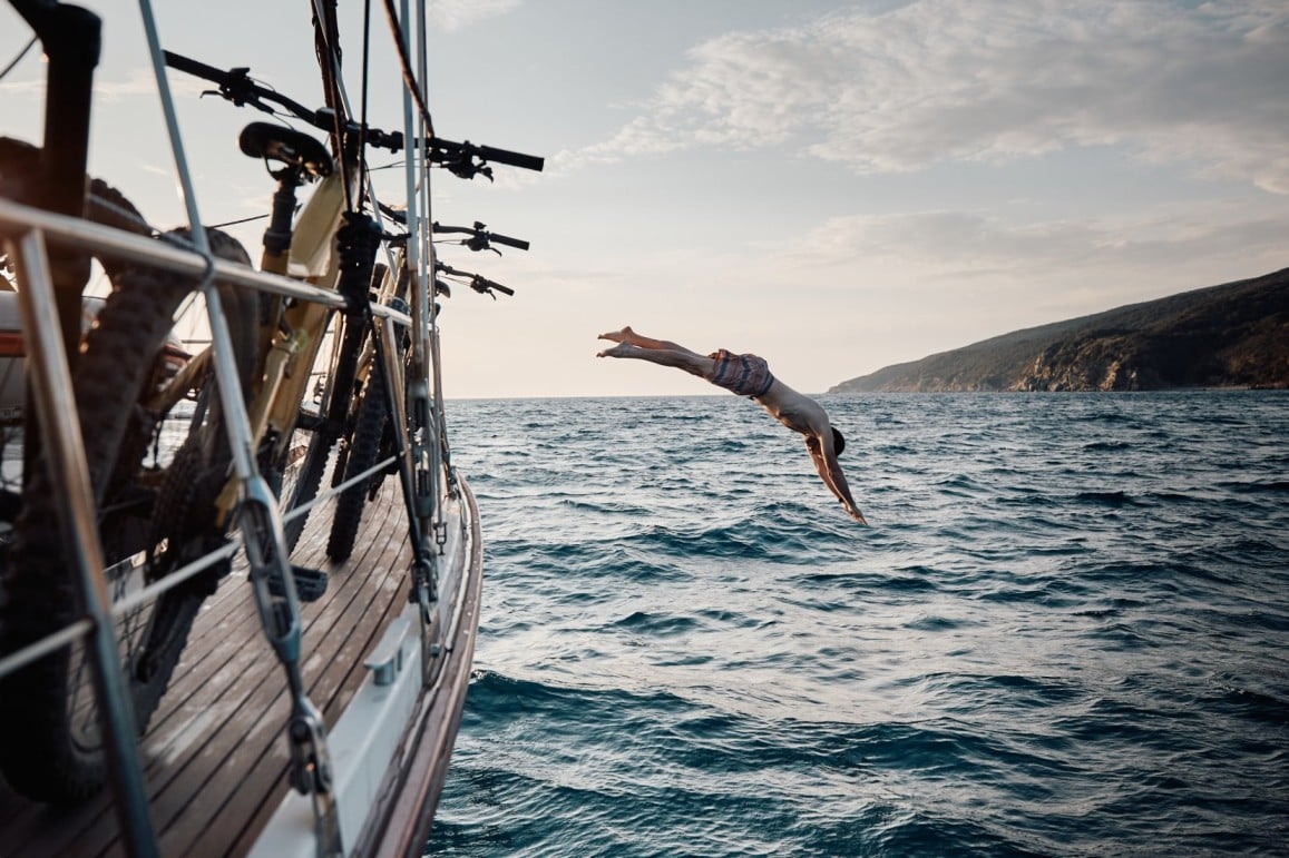 A mountain biker jumping into the sea from a sailboat with e-mountain bikes heading to Elba.