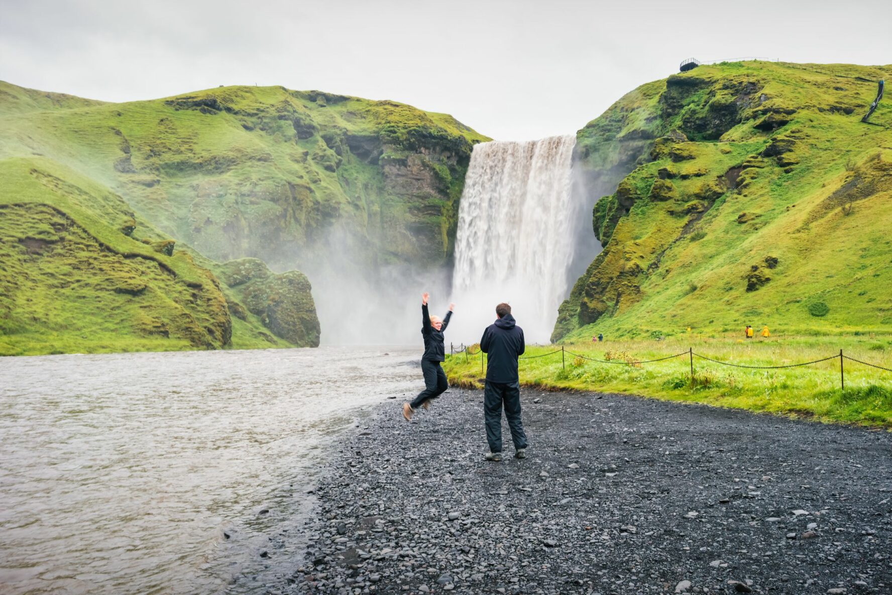 Two hikers are cheerful while appreciating the beauty of the Skogafoss waterfall in Iceland.