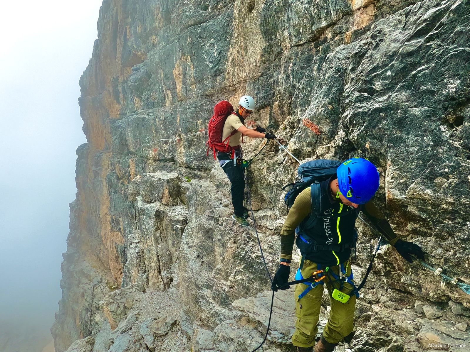 Via ferrata adventurers navigating a cabled via ferrata in the Brenta Dolomites, navigating narrow ledges of the reddish rock formations.