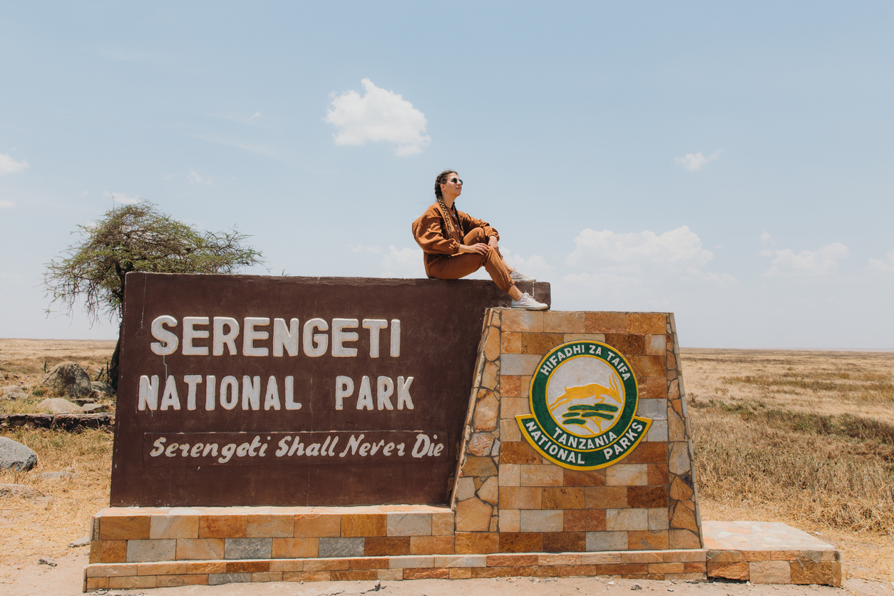 Young woman explorer in safari jumpsuit sitting on the sign contemplating the view of savannah in East Africa