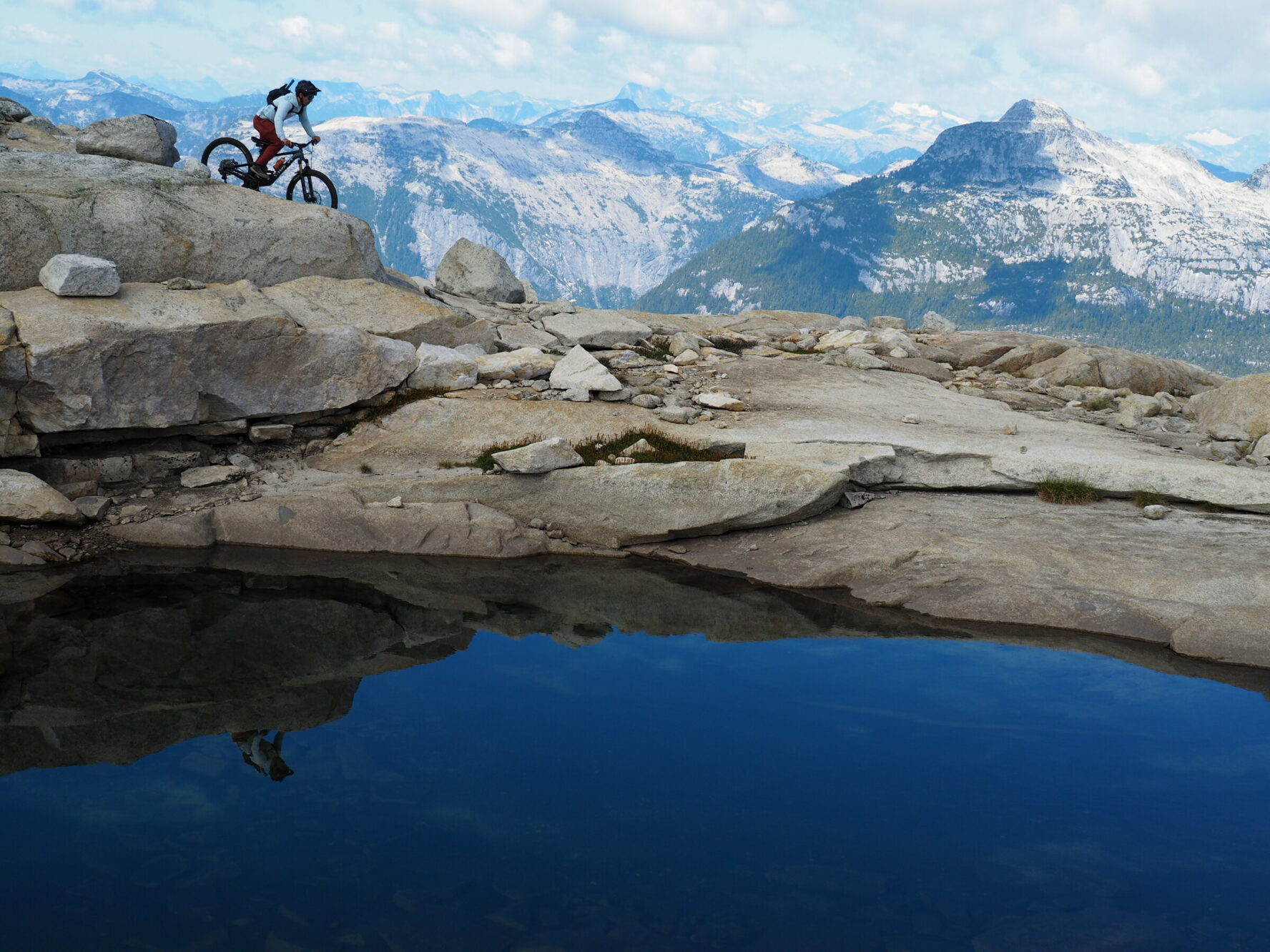 Mountain biker riding beside a small alpine lake, with high mountains in the distance.