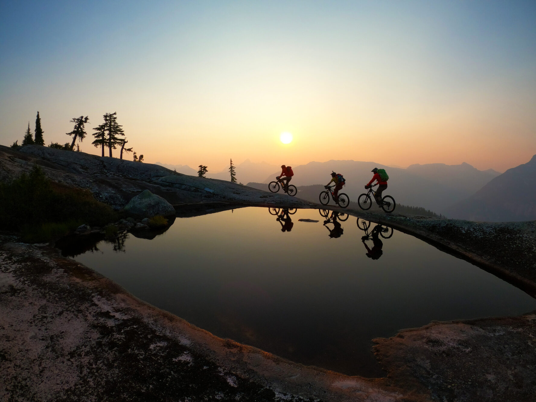 Mountain bikers’ silhouette reflecting gorgeously on the surface of a small lake in BC’s Coast Mountains in soft sunset light.
