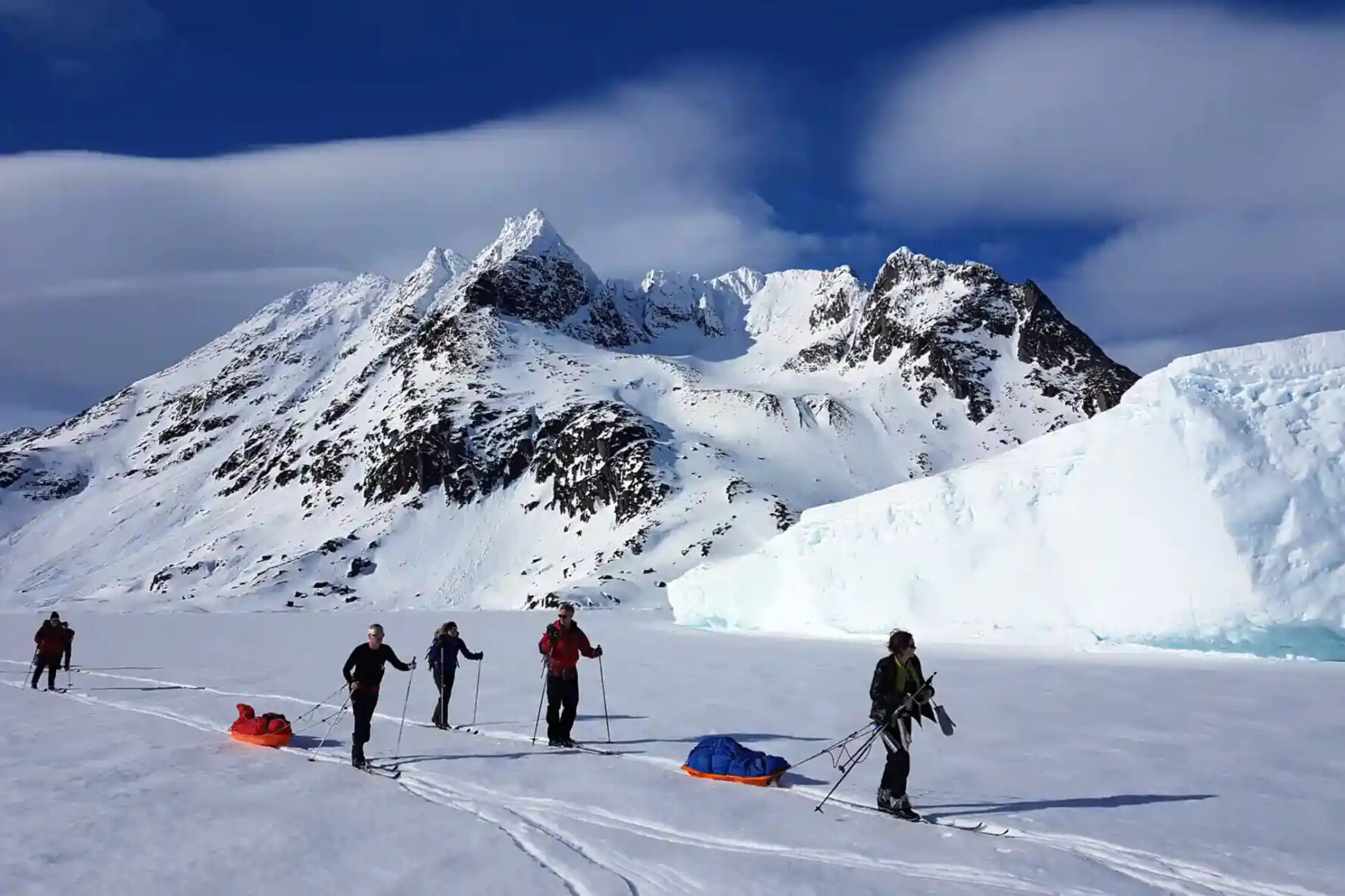 People cross-country skiing in East Greenland, with mountains and an iceberg in the background