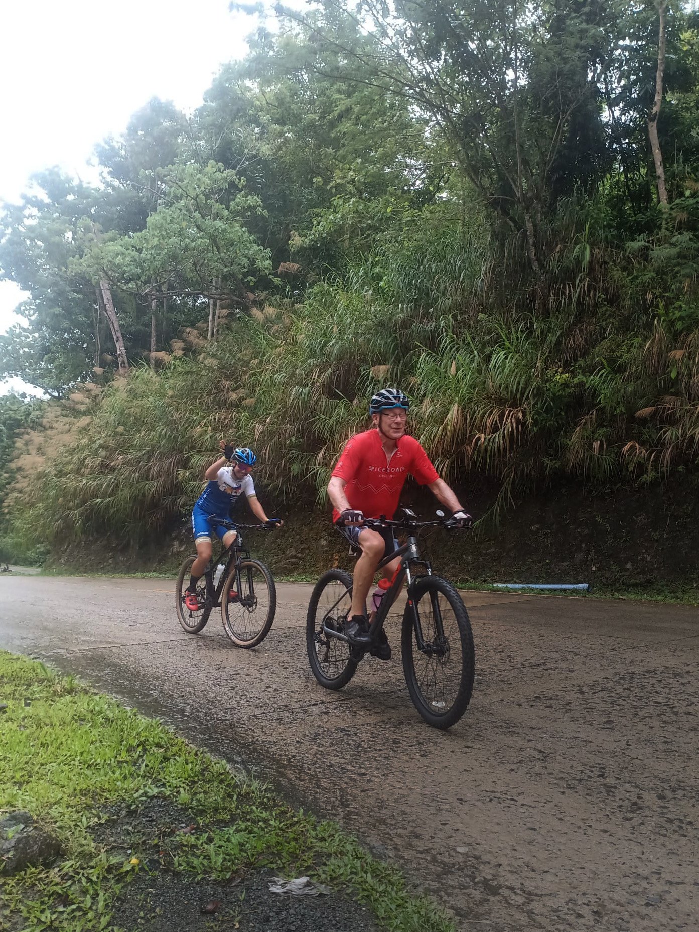 Two people cycling on a wet road surrounded by lush vegetation