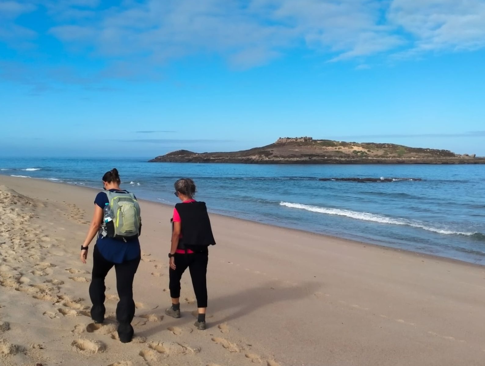 Two women walk along a sandy beach in Portugal, with a small island in the distance