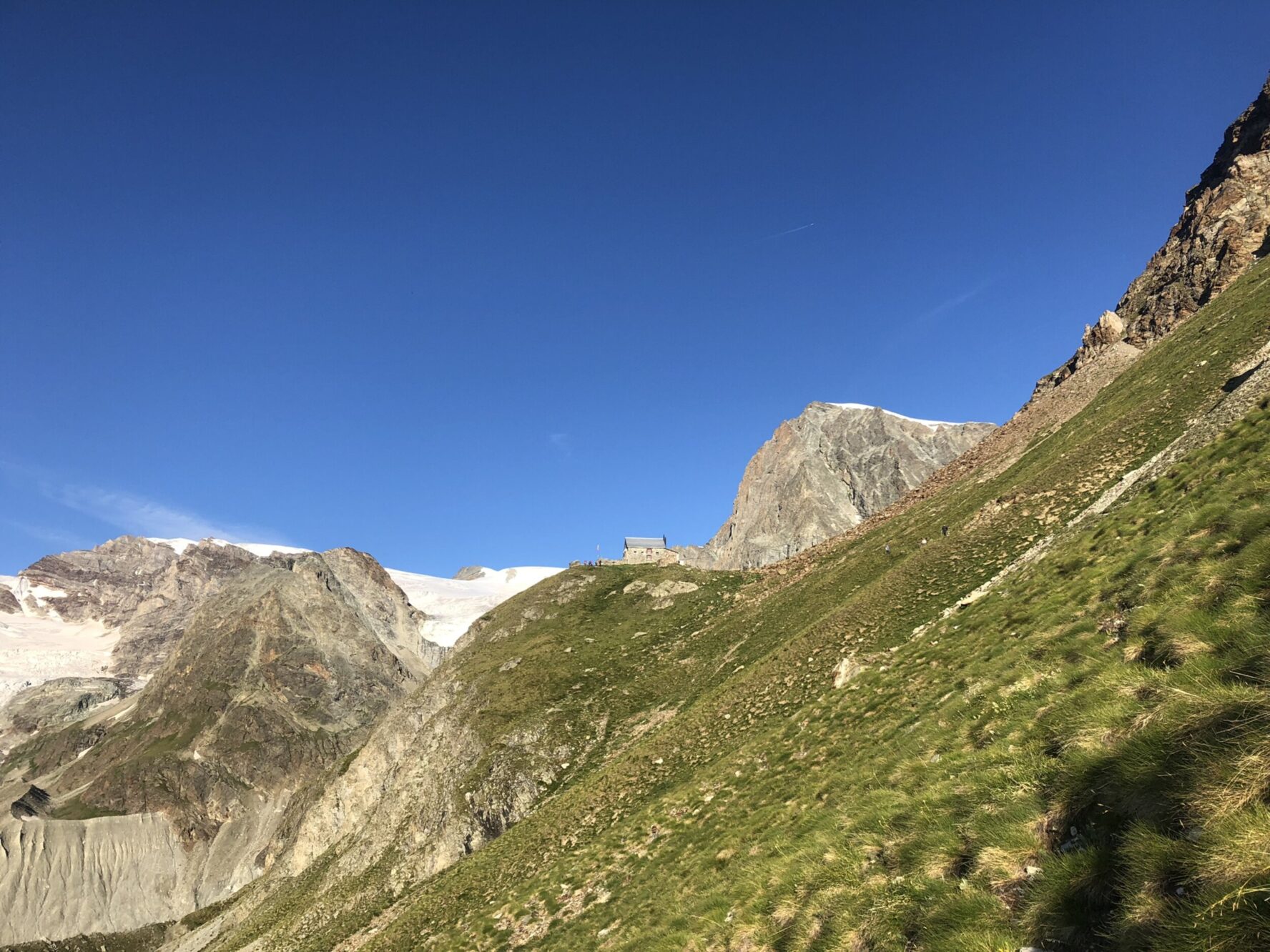 Alpine landscape with snow-capped peaks and a green slope with a hut