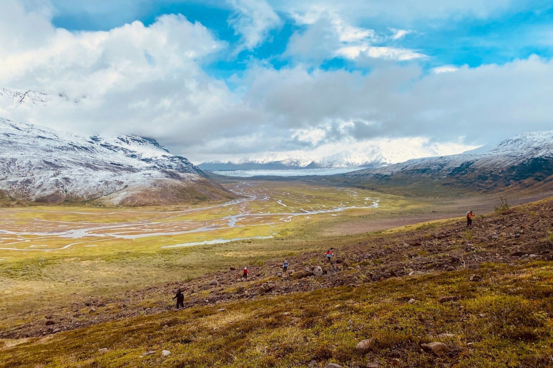 Hikers on a mountain trail overlooking a vast valley with winding glaciers and snowy peaks