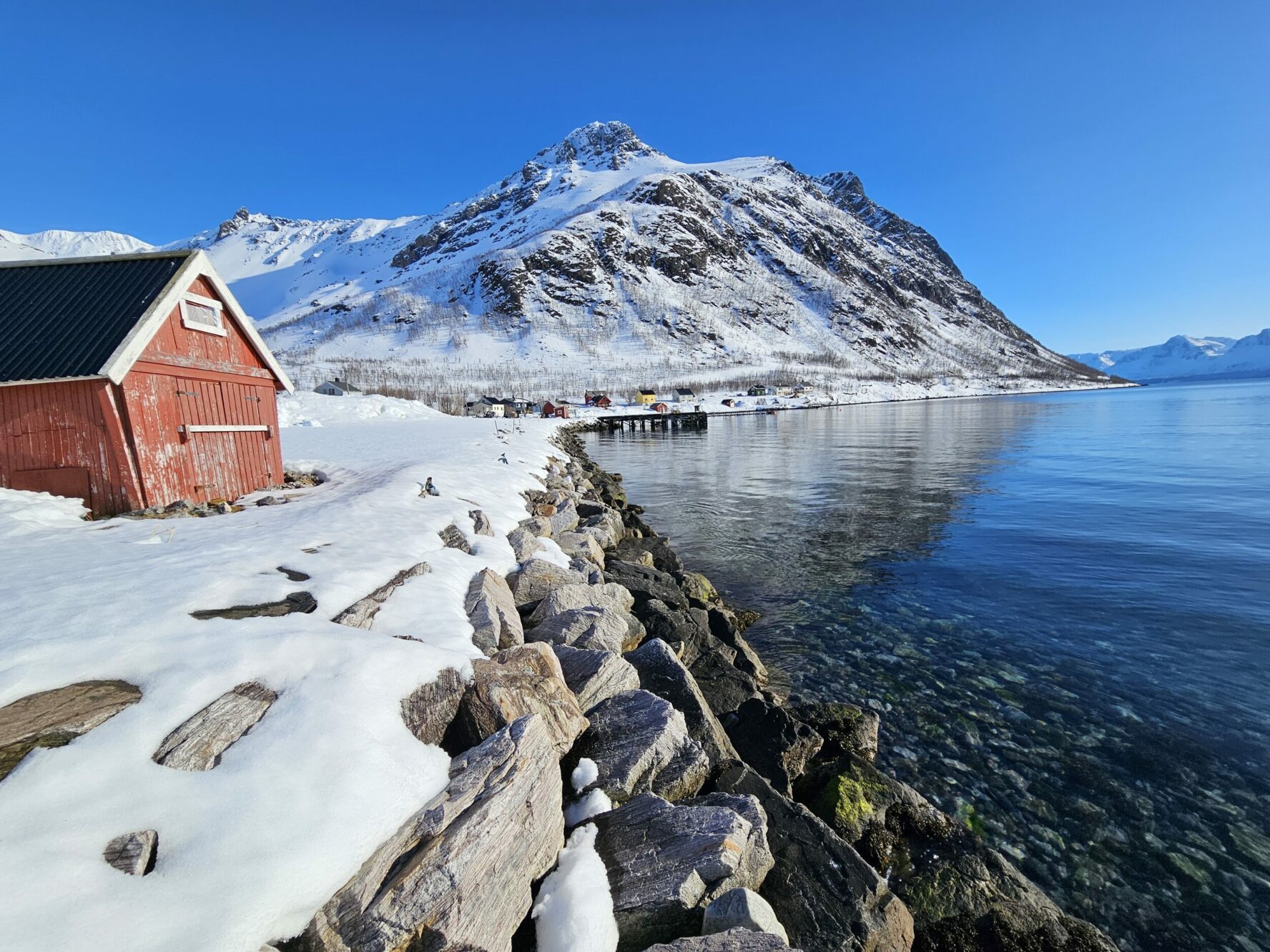A red wooden cabin by the sea with snowy mountains under a clear blue sky