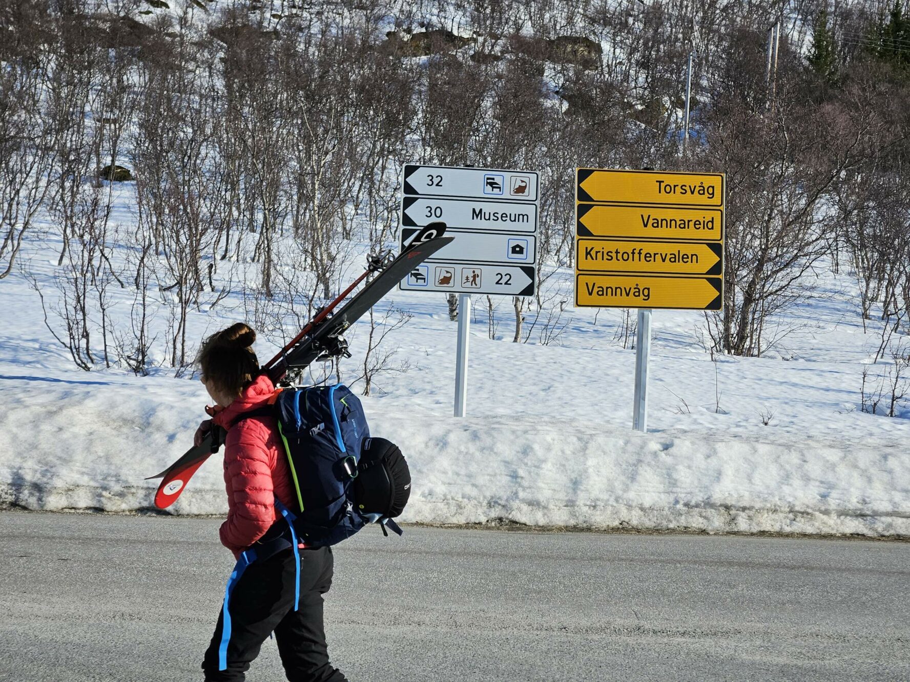 A skier walking with equipment over her shoulder on a street in Norway, with a signpost in the background