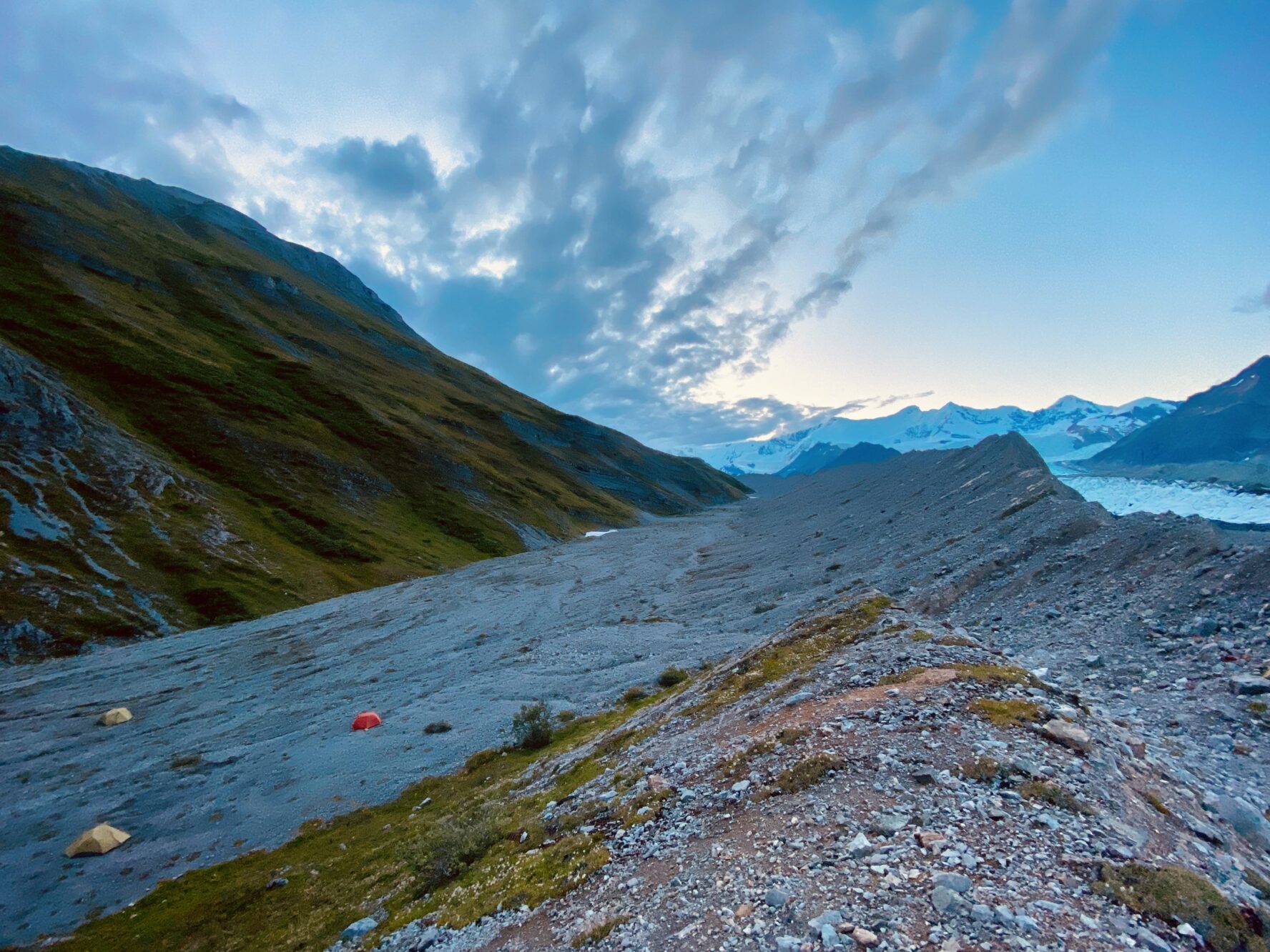 Tents in an alpine landscape at dusk with a glacier, rocky terrain, and clouds