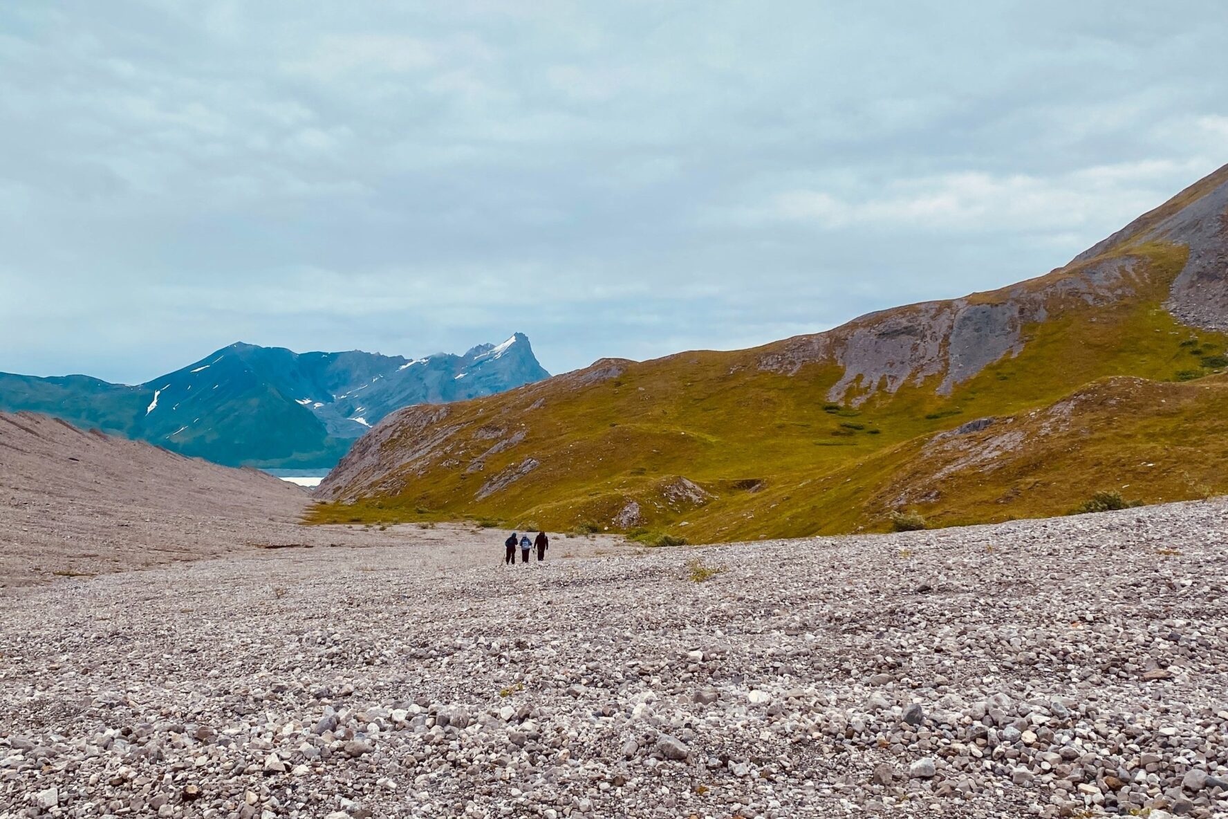 Three hikers on a rocky terrain with mountains in the background under a cloudy sky