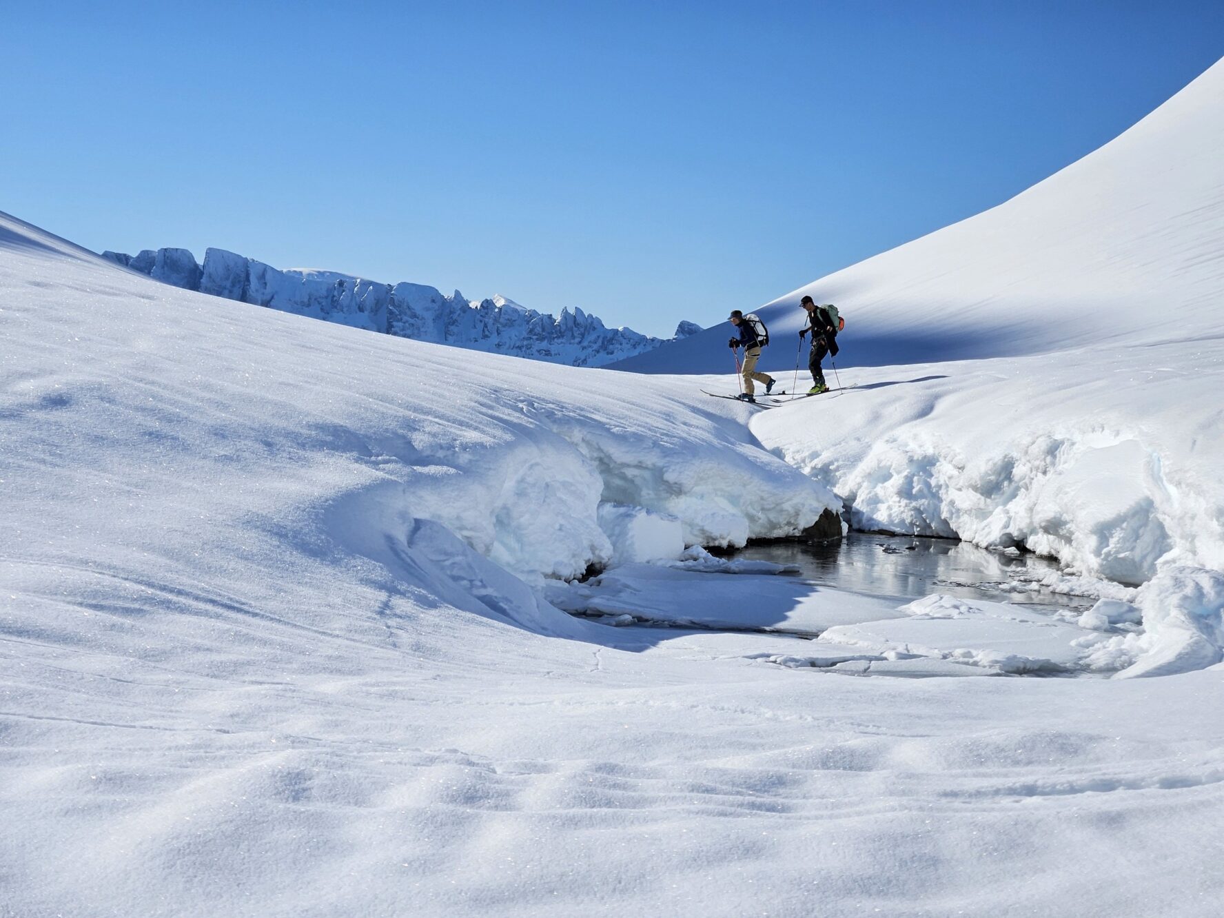 Two skiers with backpacks cross a snow-covered landscape with mountains in the background