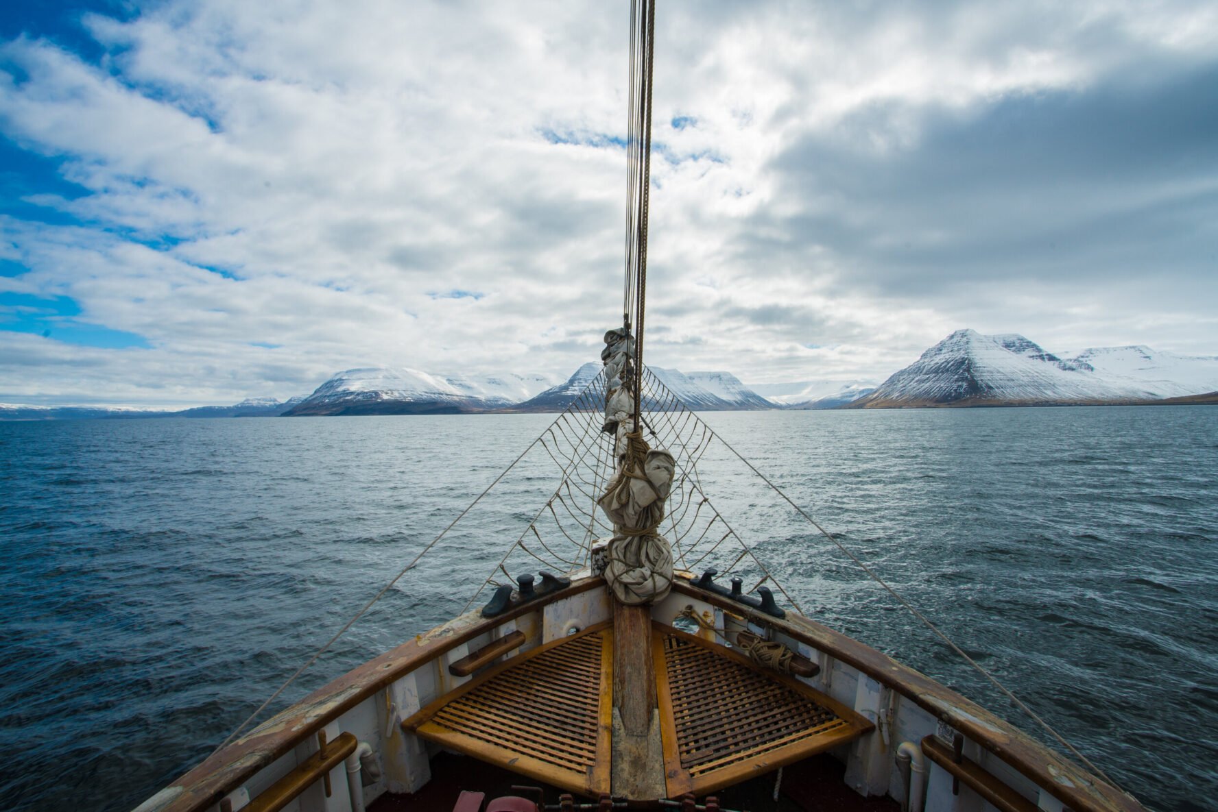 The bow of a sailboat with rigging overlooking snowy mountains and the sea