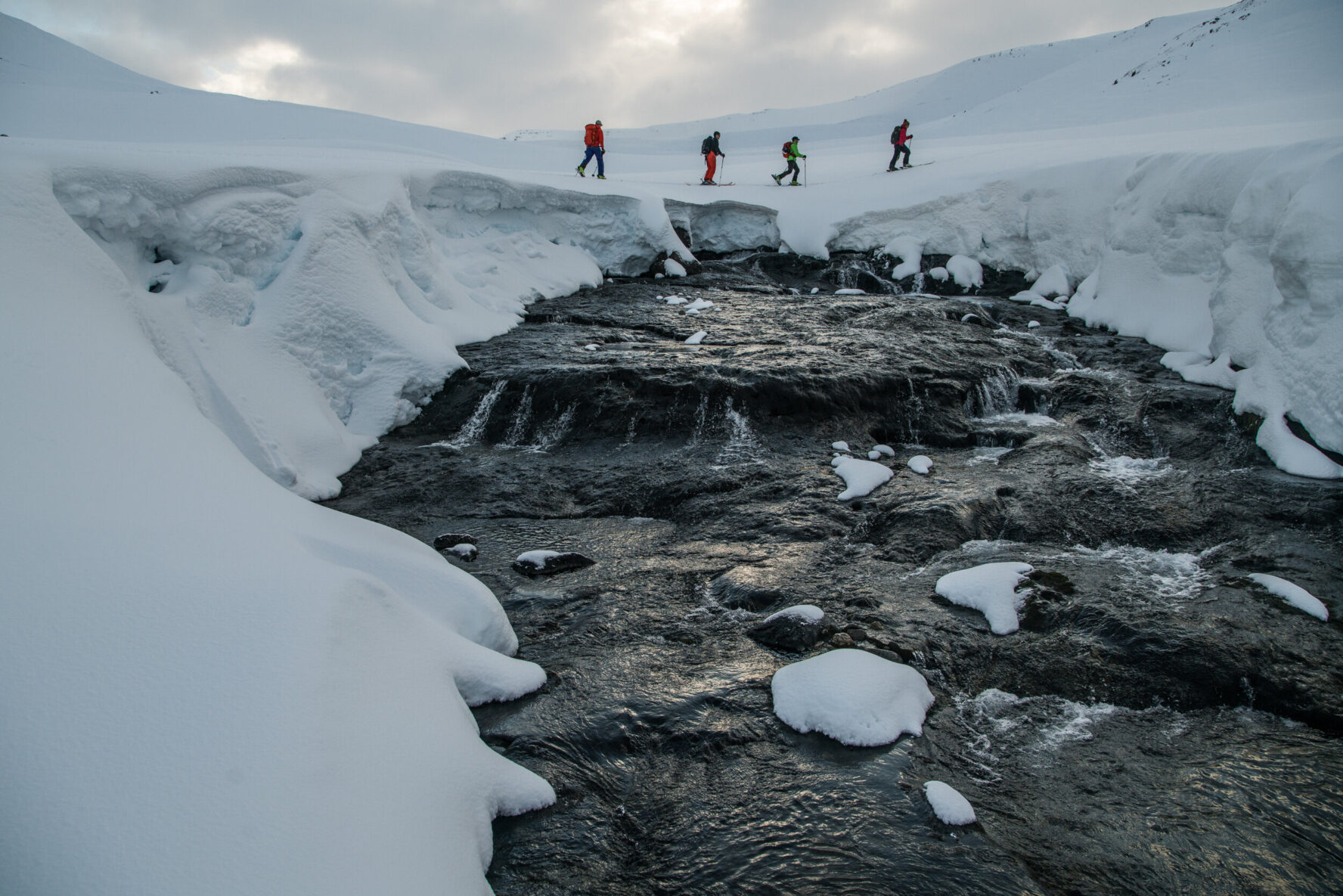 Four skiers crossing a snow-covered stream with icy banks under a cloudy sky