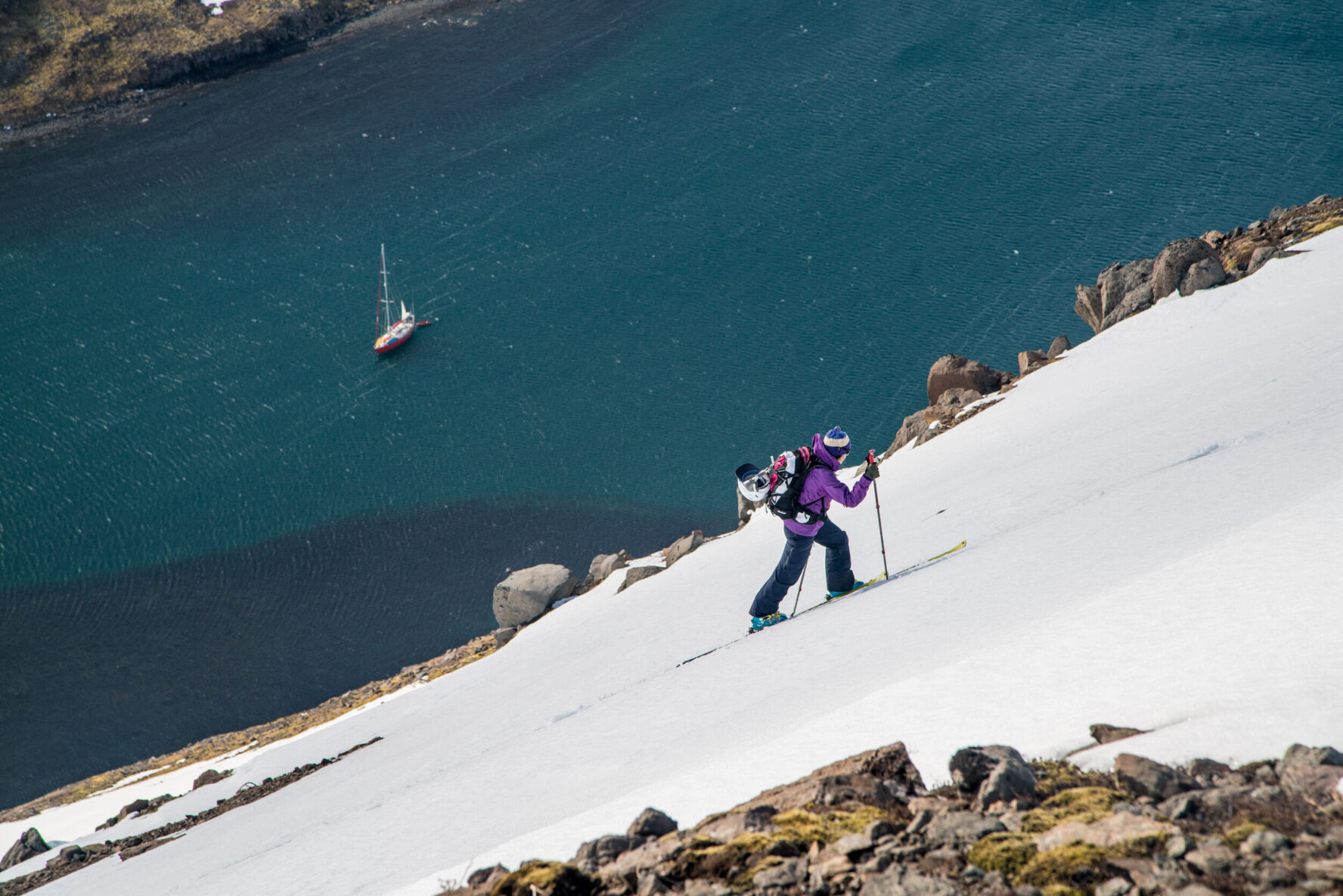 Person skiing on a snowy slope with a sailboat in the water below