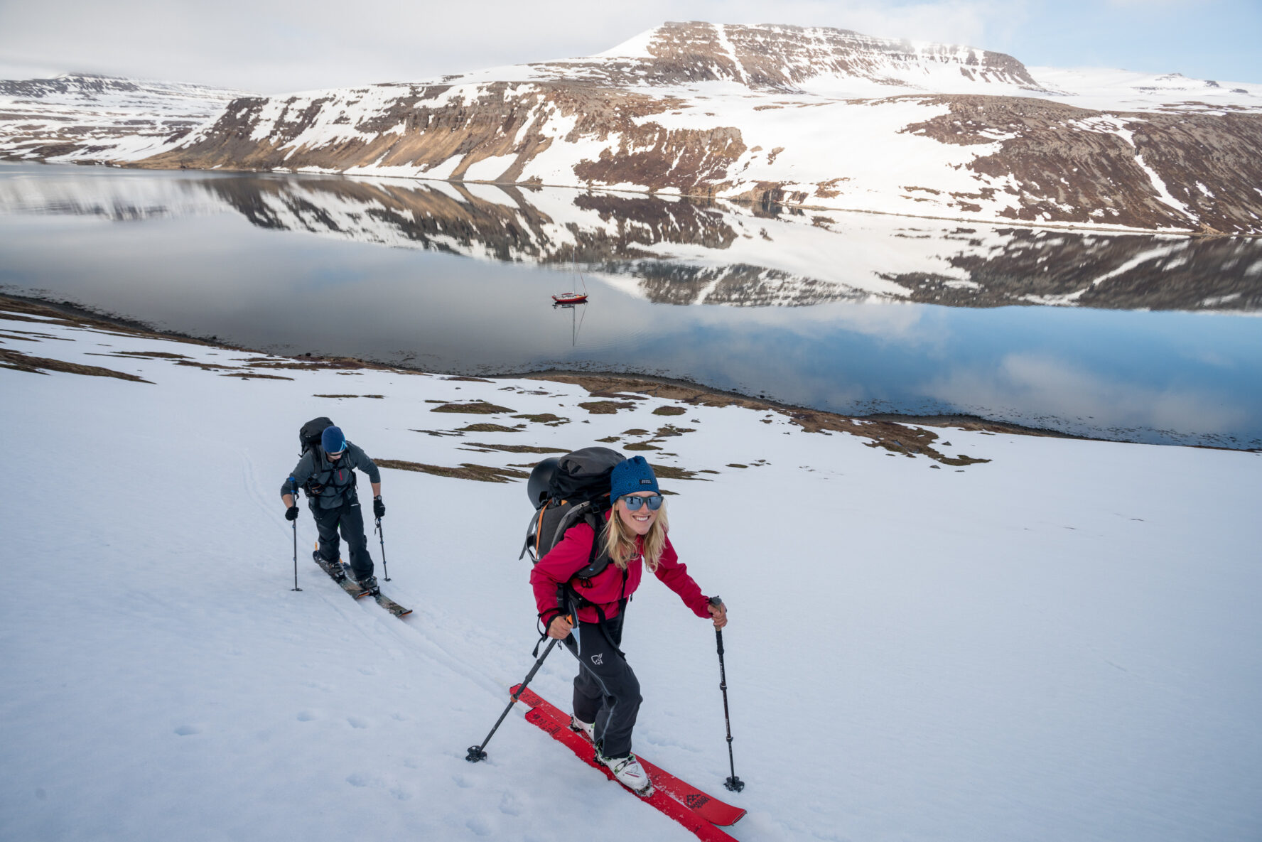 Two skiers ascending a slope with the boat, a fjord, and mountains in the background
