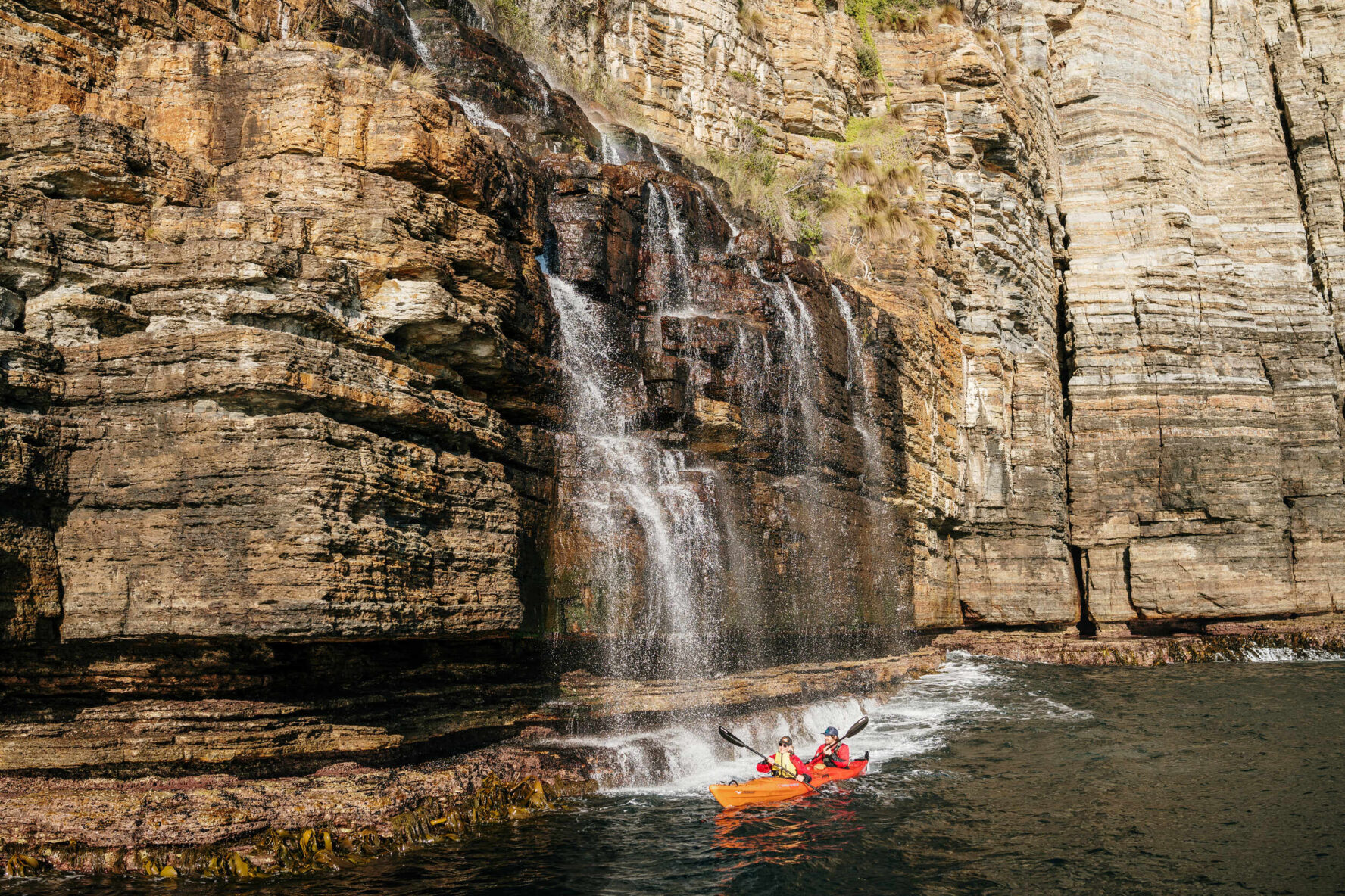 Kayakers by a cliffside waterfall in Tasmania