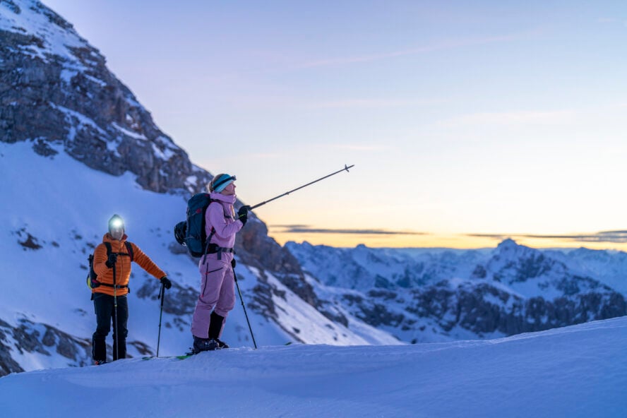 A young Caucasian couple, dressed in vibrant winter gear, ski mountaineers at sunrise in the stunning Alpine mountains. Snowy peaks and soft dawn light create a serene and adventurous atmosphere.