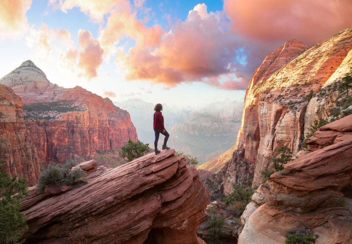 Adventurous Woman at the edge of a cliff is looking at a beautiful landscape view in the Canyon during a vibrant sunset. Taken in Zion National Park, Utah, United States.