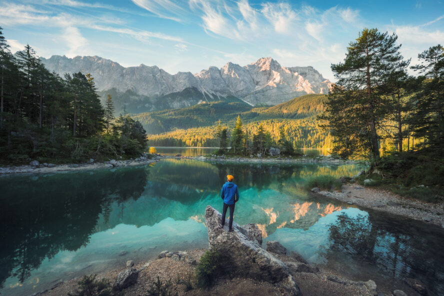 Man enjoying the amazing morning scenery at a gorgeous lake in the Bavarian Alps, with teal water reflecting the view of the mountain range and the nice clouds on a self-guided tour of the Alps