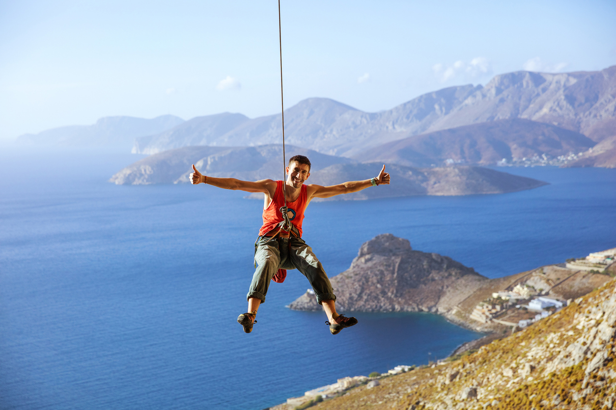 Cheerful rock climber swinging on rope and showing thumbs up against view of coast