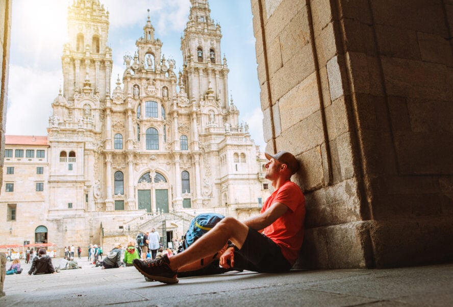 Tired middle-aged backpacker pilgrim sitting on Obradoiro Square in Santiago de Compostela, Spain, with Santiago de Compostela Cathedral, famous Camino de Santiago finish point