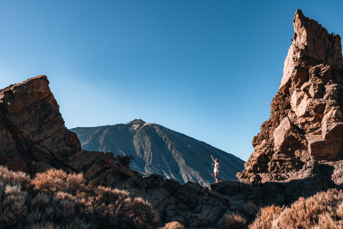 Woman hiker stands triumphantly between two large rocky formations, with a Teide mountain rising majestically in the background under a bright blue sky vista in Tenerife
