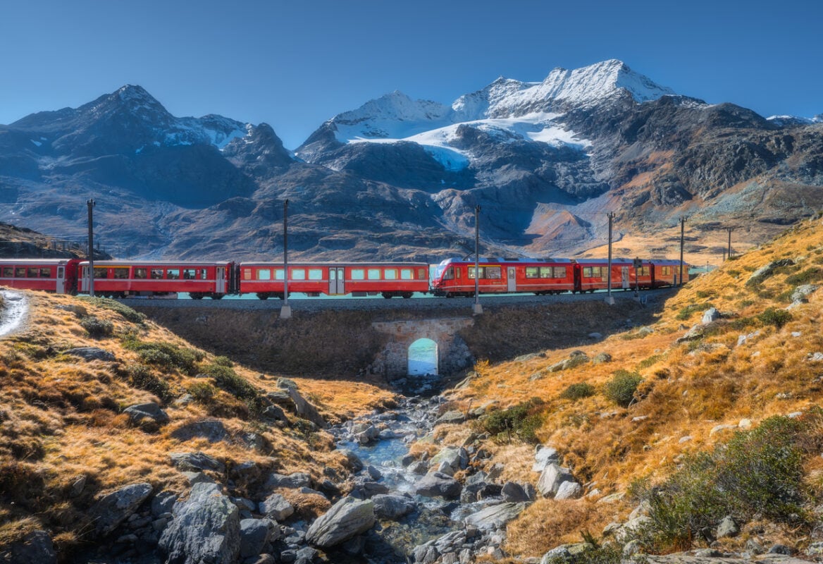 Bernina Express train crossing alpine landscape with glacier, rocky hills and clear stream in the Swiss Alps, Switzerland, on a sunny autumn day with snow-covered mountain peaks. Red train in fall
