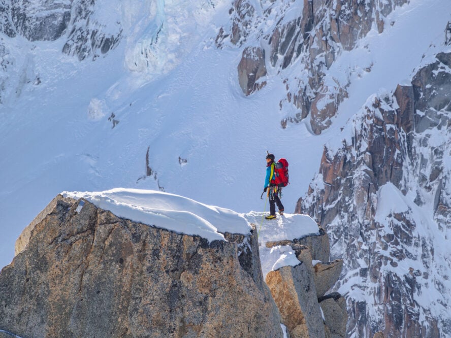 A mountaineer, fully equipped with a red backpack and ice axe, stands on a snow-covered rocky edge near Aiguille du Midi in Chamonix.